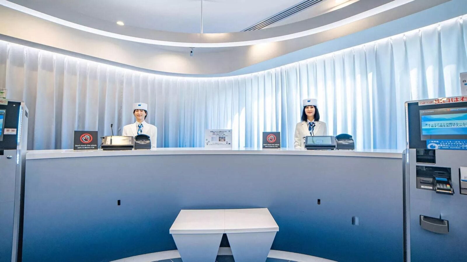 Dwa humanoidalne roboty stoją za biurkiem recepcji hotelu obsługiwanego przez roboty