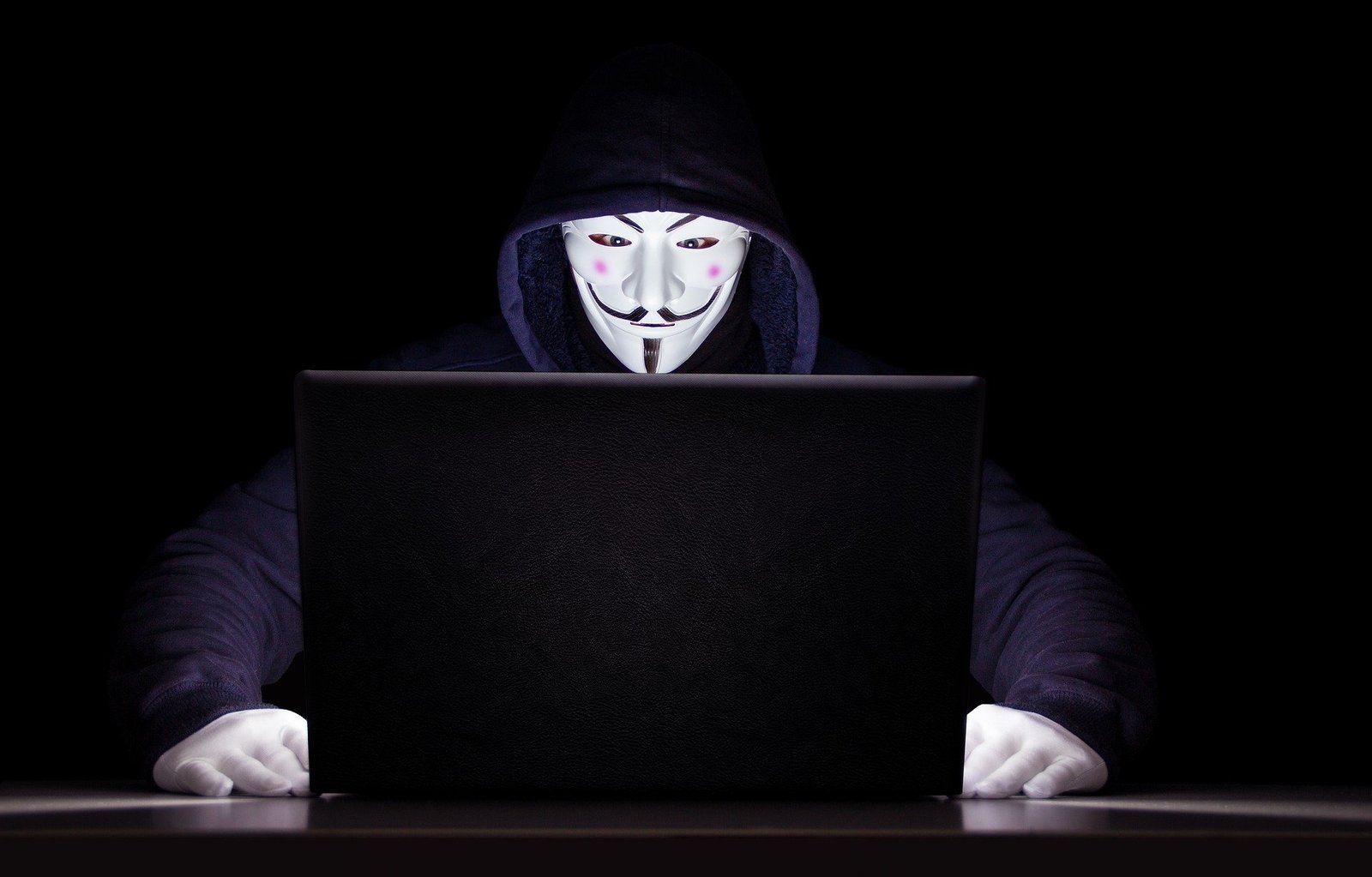 Haker w masce siedzący przed czarnym laptopem